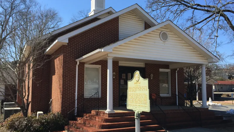 Historic brick building with white columns and a historical marker sign in front under clear blue sky