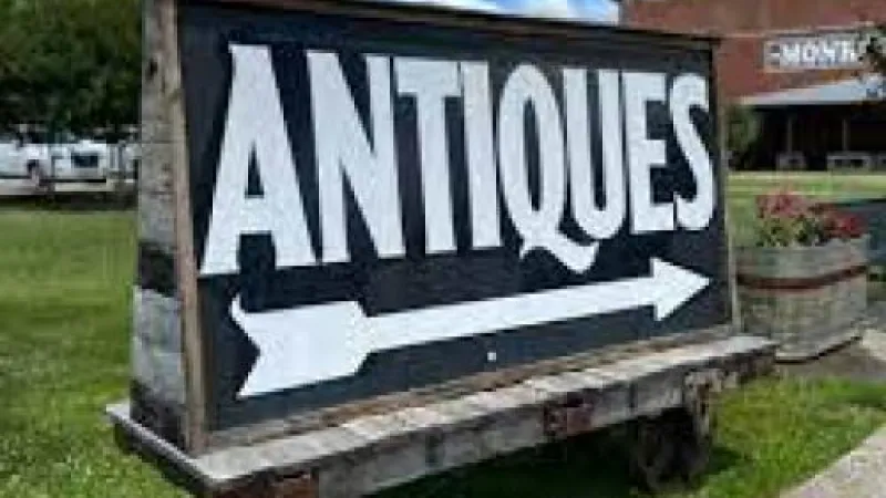 Large wooden sign with white ANTiques text and arrow pointing right on grass near a brick building under blue sky.