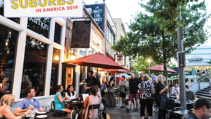 Outdoor dining and socializing on a lively suburban street with trees, umbrellas, and restaurants at dusk.