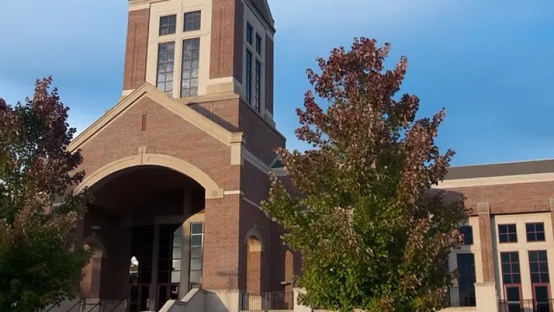 Brick church building with tall bell tower and cross under clear blue sky with trees in front.