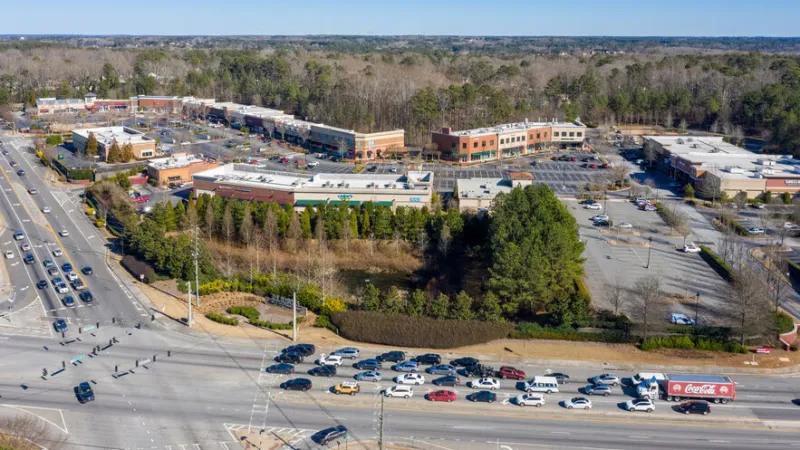 Aerial view of suburban shopping center with parked cars, adjacent forest, and busy multi-lane road with vehicles.