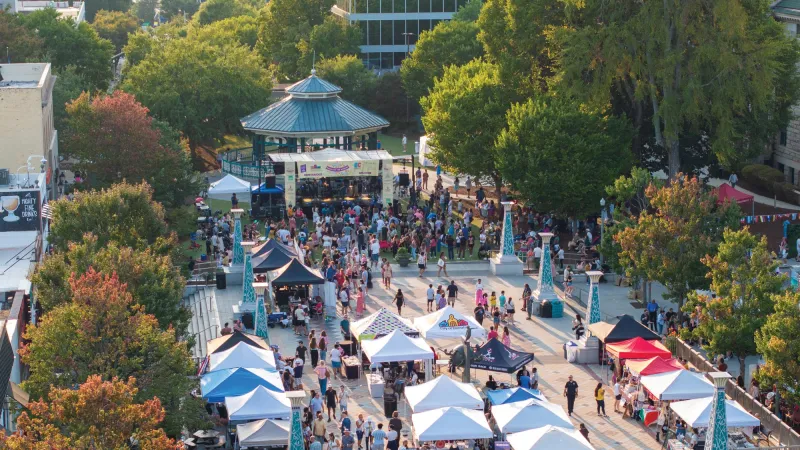 Aerial view of a crowded outdoor festival with tents, a stage, and people enjoying the event surrounded by trees.