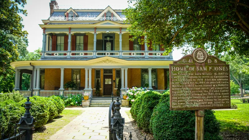 Historic two-story yellow home with porch, gardens, and a Sam Jones historical marker in front under tree shade.