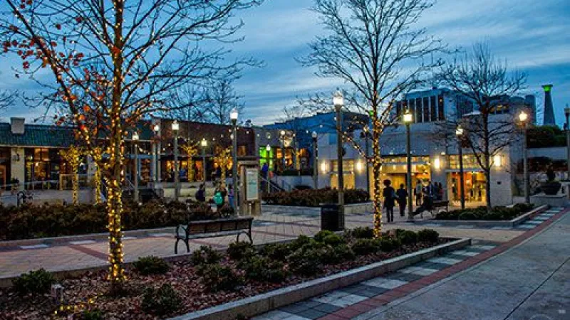 Evening city plaza with lit trees, benches, people, and buildings under a cloudy blue sky at dusk.