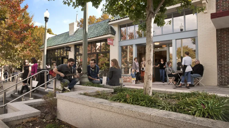 People enjoying a sunny day outside a bakery with outdoor seating and live guitar music on a tree-lined street.