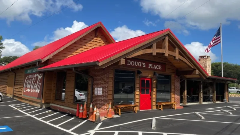 Exterior of Doug's Place restaurant with red roof, wooden walls, red door, Coca-Cola sign, and American flag.