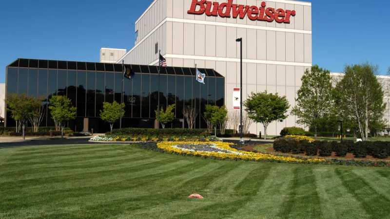 Budweiser brewery building with large red logo, surrounded by green lawn, flowers, and trees under blue sky