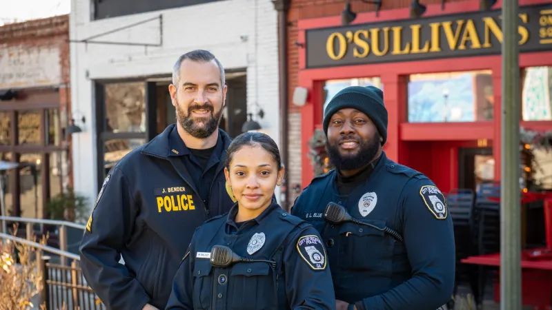 Three police officers smiling outdoors in uniform in front of O'Sullivans restaurant on a sunny day.