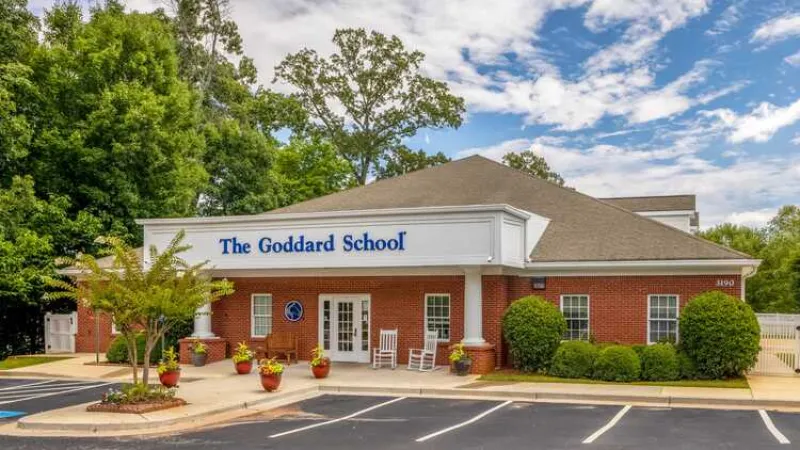 Brick building of The Goddard School with white columns and rocking chairs at entrance under a partly cloudy sky.