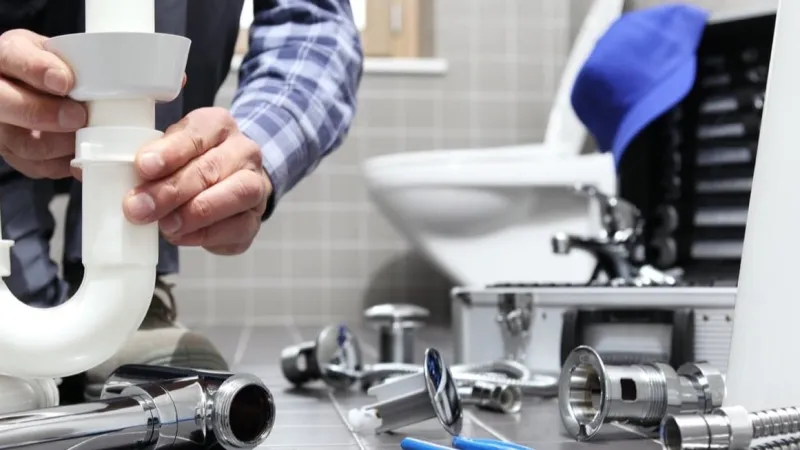 Plumber assembling white PVC pipe under a sink amid plumbing tools and parts in a tiled bathroom.