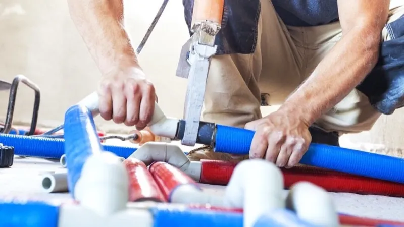 Plumber working with blue and red pipes using a cutting tool on a concrete floor in construction site.