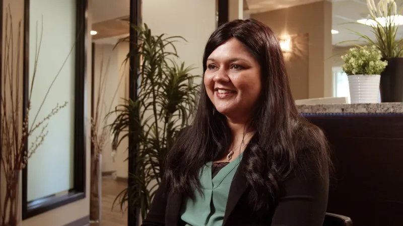 Smiling woman with long dark hair sitting indoors near plants and modern office decor, speaking or listening attentively