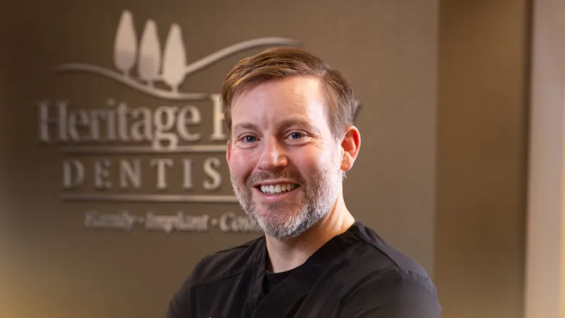 Smiling male dentist Dr. Bradshaw in black scrubs posing in front of Heritage Family Dentistry sign.