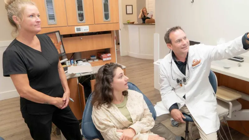 Dentist in white coat explains treatment to female patient in dental office while assistant observes.