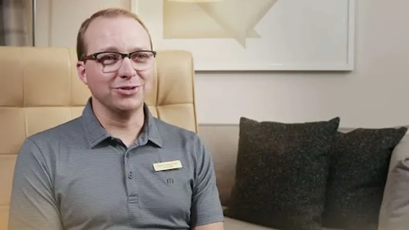 Smiling man in glasses and gray polo shirt sitting in a beige chair with dark cushions and wall art behind.