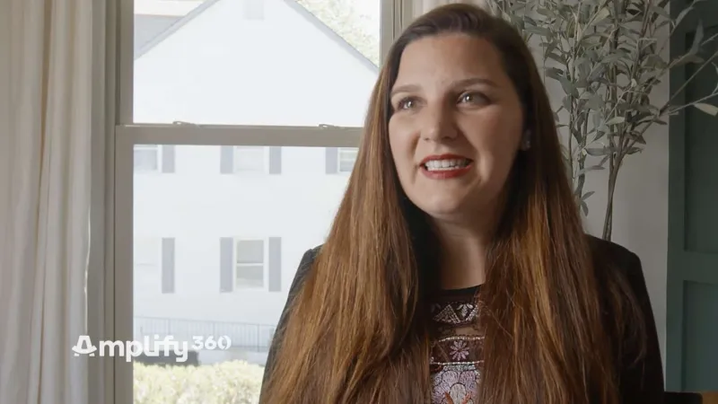 Smiling woman with long brown hair standing near window in bright room with house visible outside.