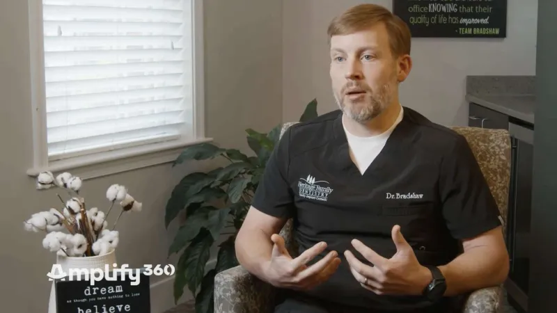 Male doctor in black scrubs speaking during interview in a well-lit room with plants and inspirational decor.
