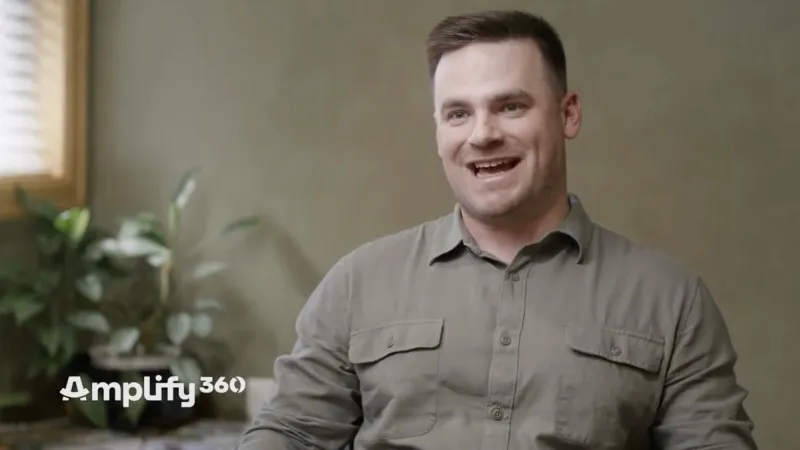 Man in casual shirt smiling and speaking in a well-lit room with indoor plants in the background.