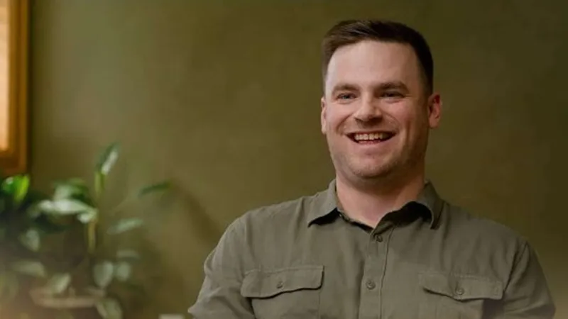 Smiling man in green shirt sitting indoors with a blurred plant and window background