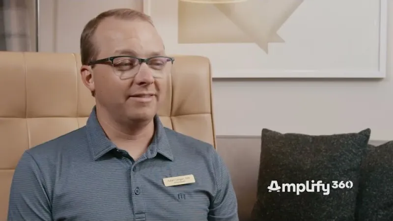 Man with glasses and name tag sitting in beige chair with gray pillows and Amplify360 logo in background.