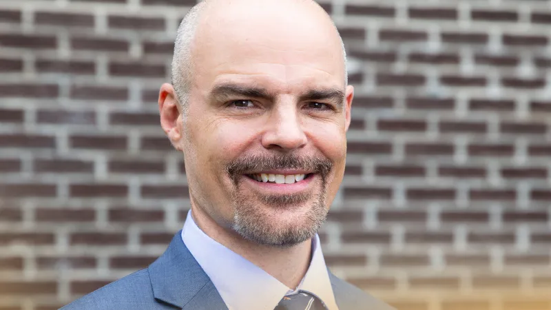 Smiling bald man with a goatee wearing a blue suit and tie standing in front of a brick wall outdoors