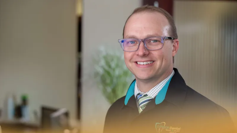 Smiling man in glasses wearing a dental coat with London Dental logo in a softly lit office background.