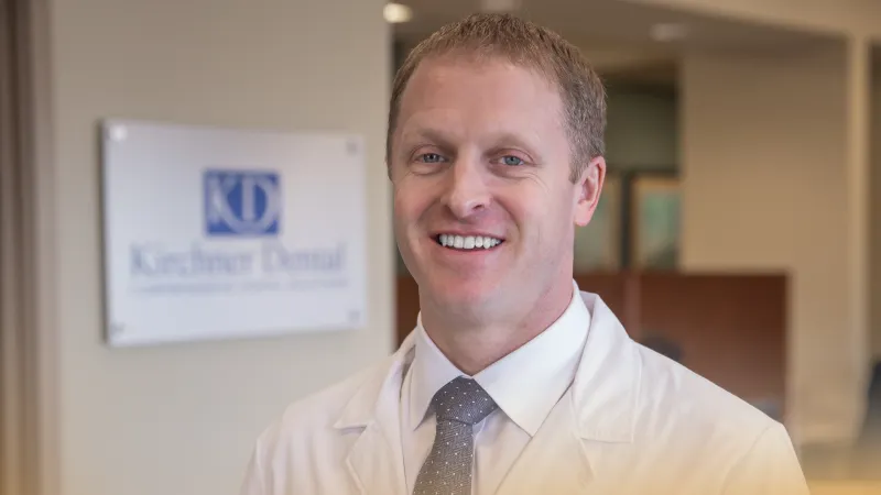 Smiling male dentist in white coat standing in dental office with Kiechner Dental sign in background