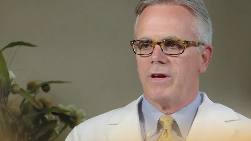 Middle-aged man in white coat and glasses speaking indoors with plants in the background.