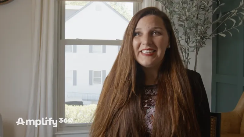 Smiling woman with long brown hair standing near window in bright room with house visible outside.