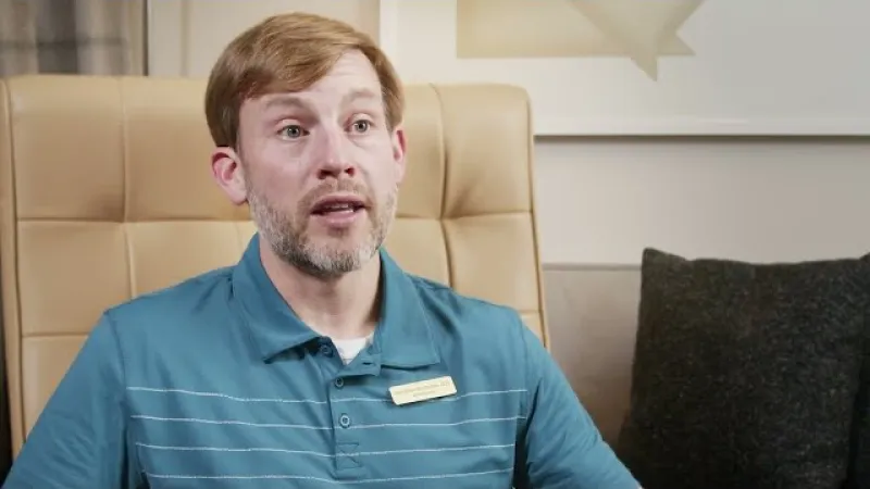 Man with beard wearing blue polo shirt sitting on beige chair and speaking indoors with grey pillow in background
