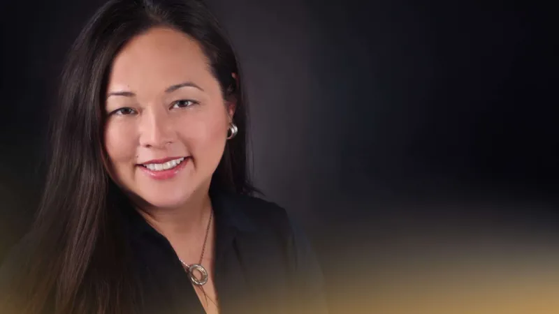 Smiling woman with long dark hair wearing a black top and silver necklace against a dark background