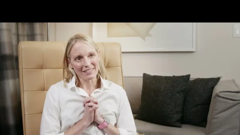 Smiling woman in white shirt sitting on tan chair with gray sofa and pillows in background indoors.