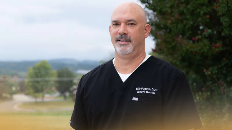 Smiling male dentist in black scrubs standing outdoors with trees and a blurred scenic background.