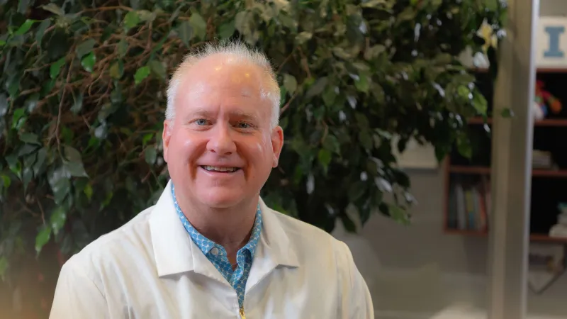 Smiling middle-aged man in a white lab coat with a blue shirt sits indoors with plants and bookshelves in the background.