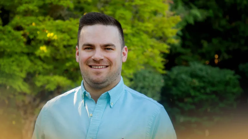 Smiling man in light blue button-up shirt standing outdoors with green foliage in the background.