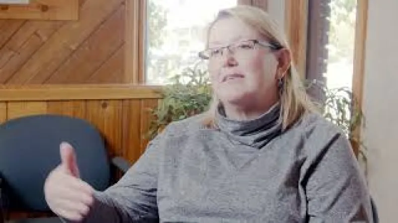 Woman in gray sweater and glasses speaking and gesturing with thumb up in a wood-paneled room with window.