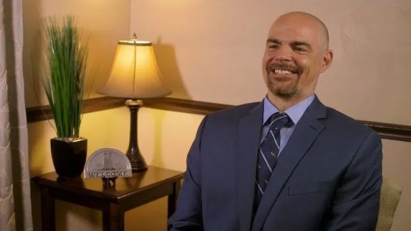 Smiling bald man in blue suit and striped tie sitting by lamp and plant in cozy room with warm lighting