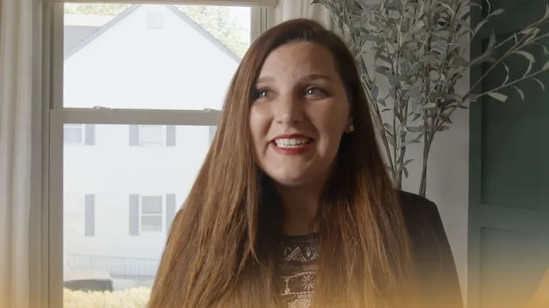Smiling woman with long brown hair standing indoors near a window with white curtains and green wall plants.