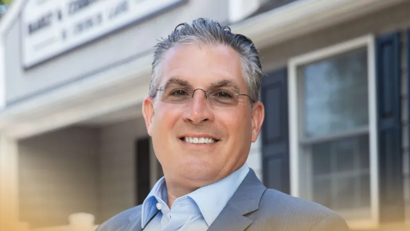 Smiling middle-aged man in glasses and suit jacket standing outdoors near a building with windows.