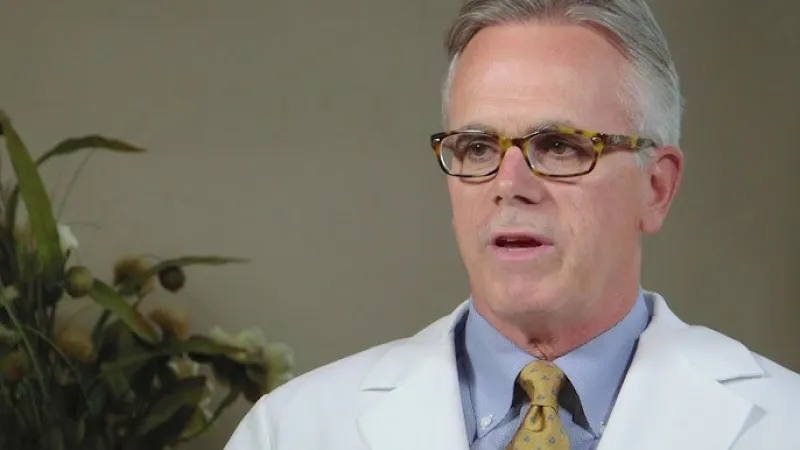 Mature male doctor wearing glasses, white coat, yellow tie, speaking in a professional setting with plant background