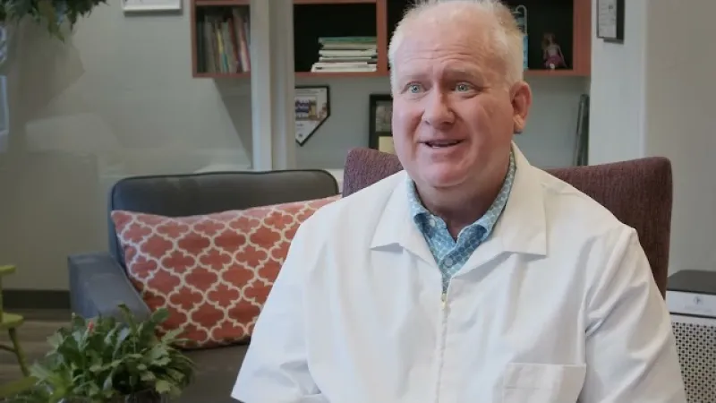 Middle-aged man in white lab coat sitting in office with bookshelf and patterned pillow behind him.