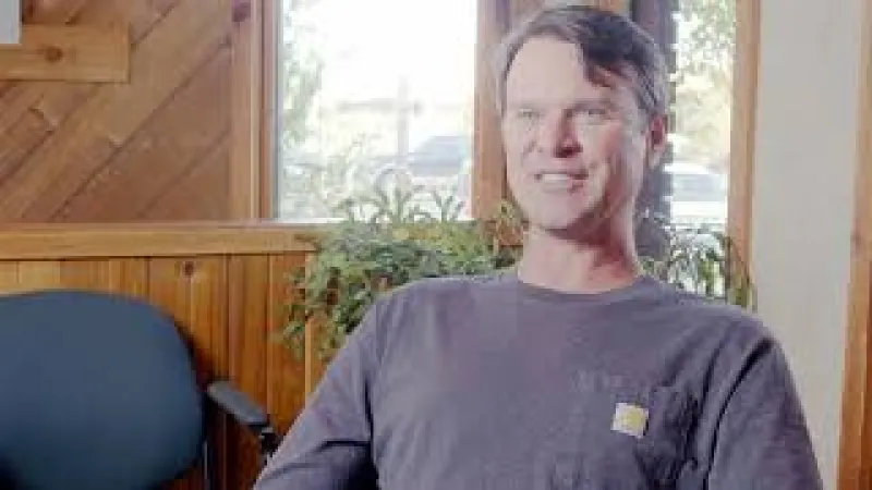 Man in casual shirt sitting in a wooden room with plants and natural light from a window behind him