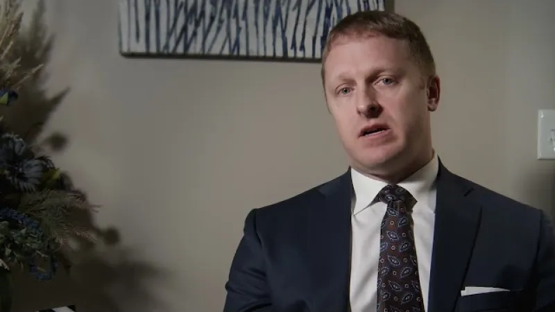 Man in dark suit and patterned tie sitting indoors with neutral background and decorative plants.