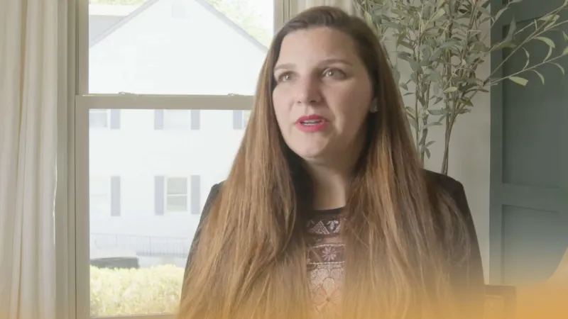 Woman with long brown hair speaking indoors next to a window with a white house outside