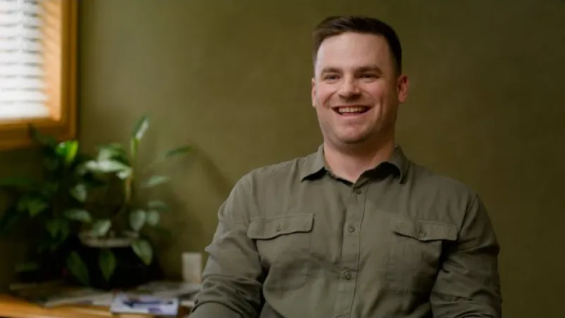 Smiling man in olive green shirt sitting indoors with plants and window light in background.