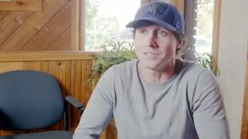 Man wearing a blue cap and gray long-sleeve shirt sitting indoors with wooden walls and a window in the background