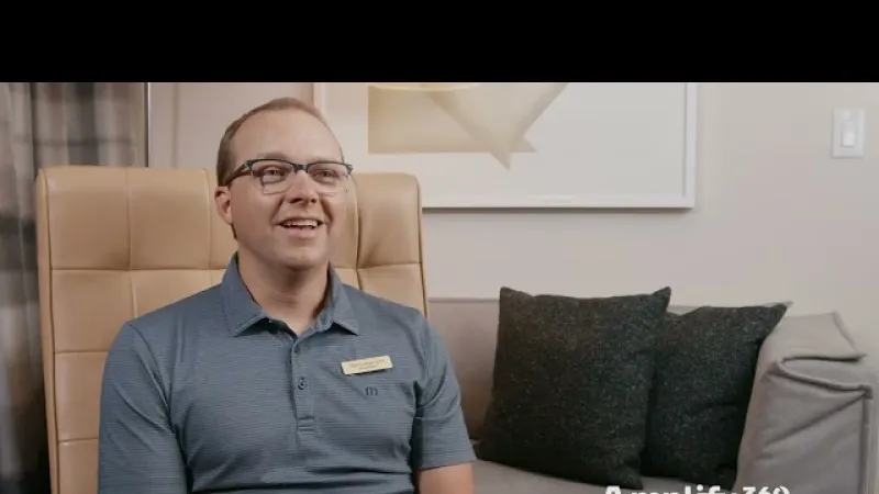 Smiling man wearing glasses and blue polo sits in beige chair next to sofa with black pillows in living room
