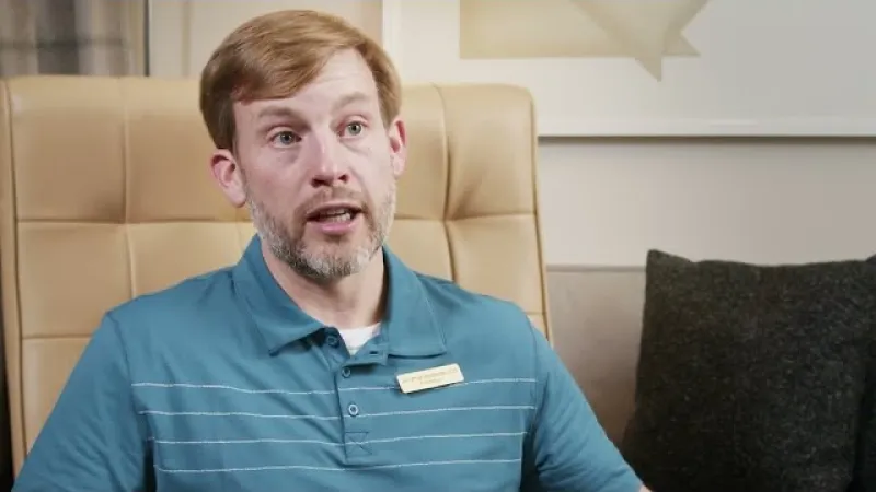 Man with light brown hair and beard wearing a teal polo shirt sitting on a beige chair talking indoors.