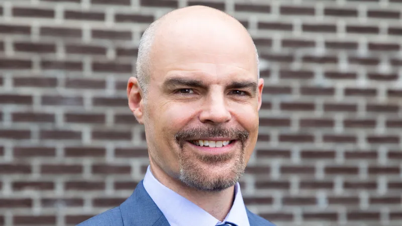Smiling bald man in blue suit and patterned tie standing in front of a brick wall outdoors.