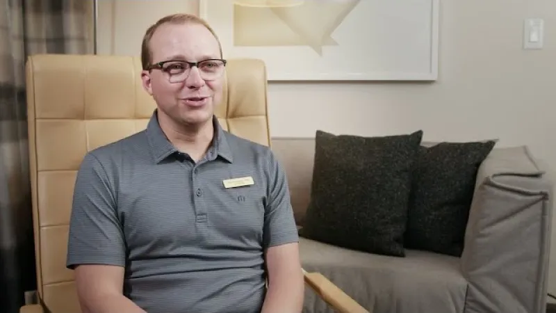Man wearing glasses and grey polo shirt sitting on a beige chair in a modern living room setting.